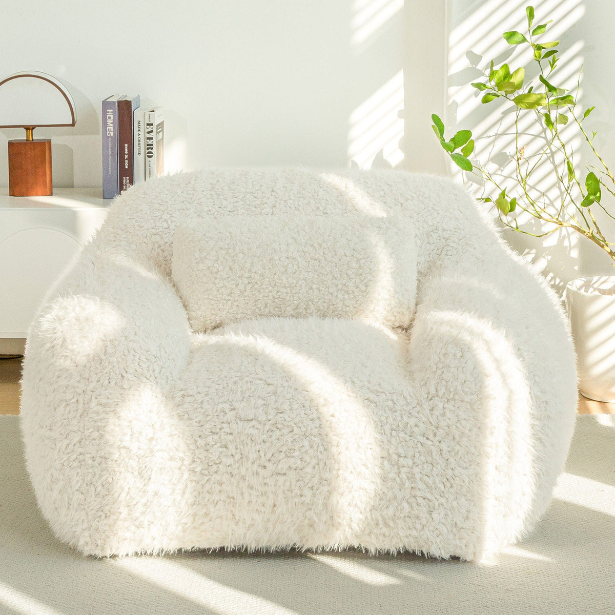 A large beige fluffy bean bag chair with a pillow, placed in a bright room with natural lighting and some plants.